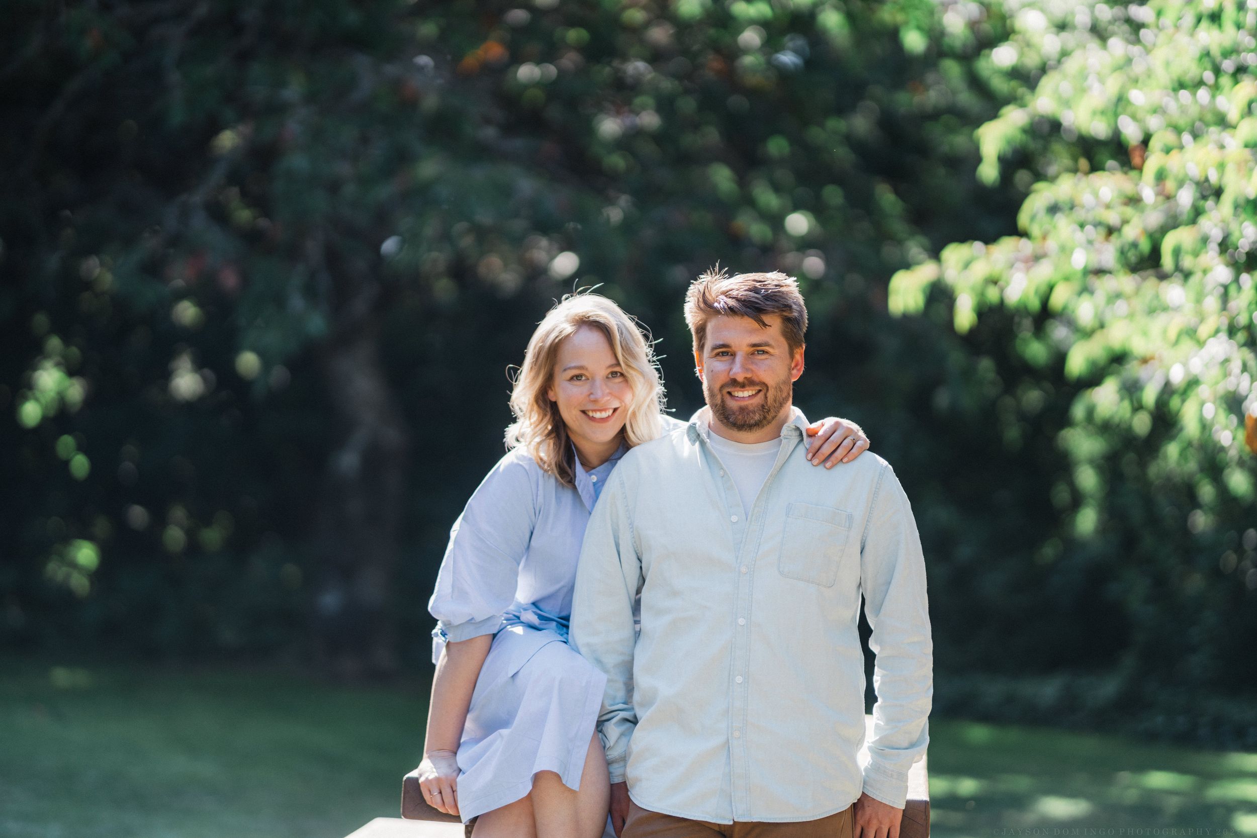A casual couple poses outdoors together wearing light colored clothing against a green garden background.