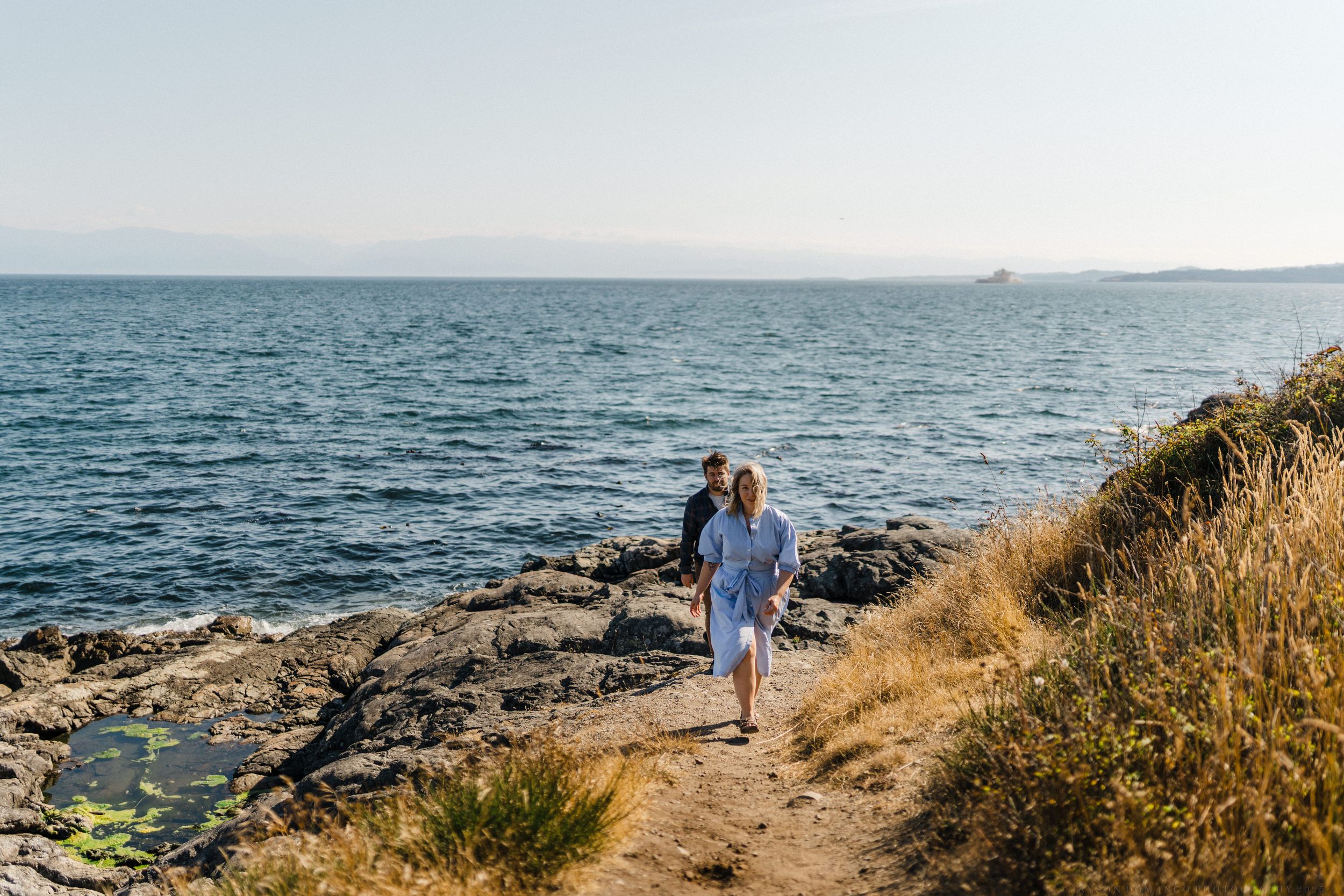 Couple walking on a coastal hiking trail alongside blue ocean waters and rocky cliffs on a sunny day.