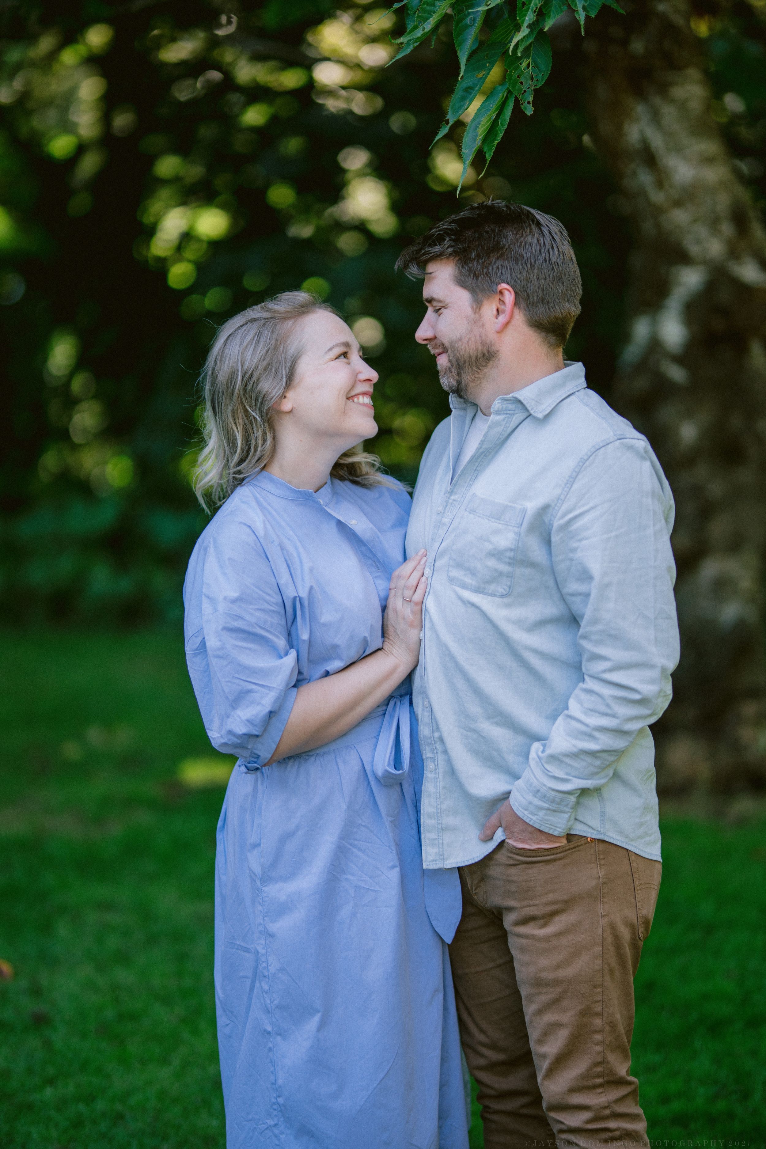 Couple sharing loving moments in blue dress and white shirt against lush green background.