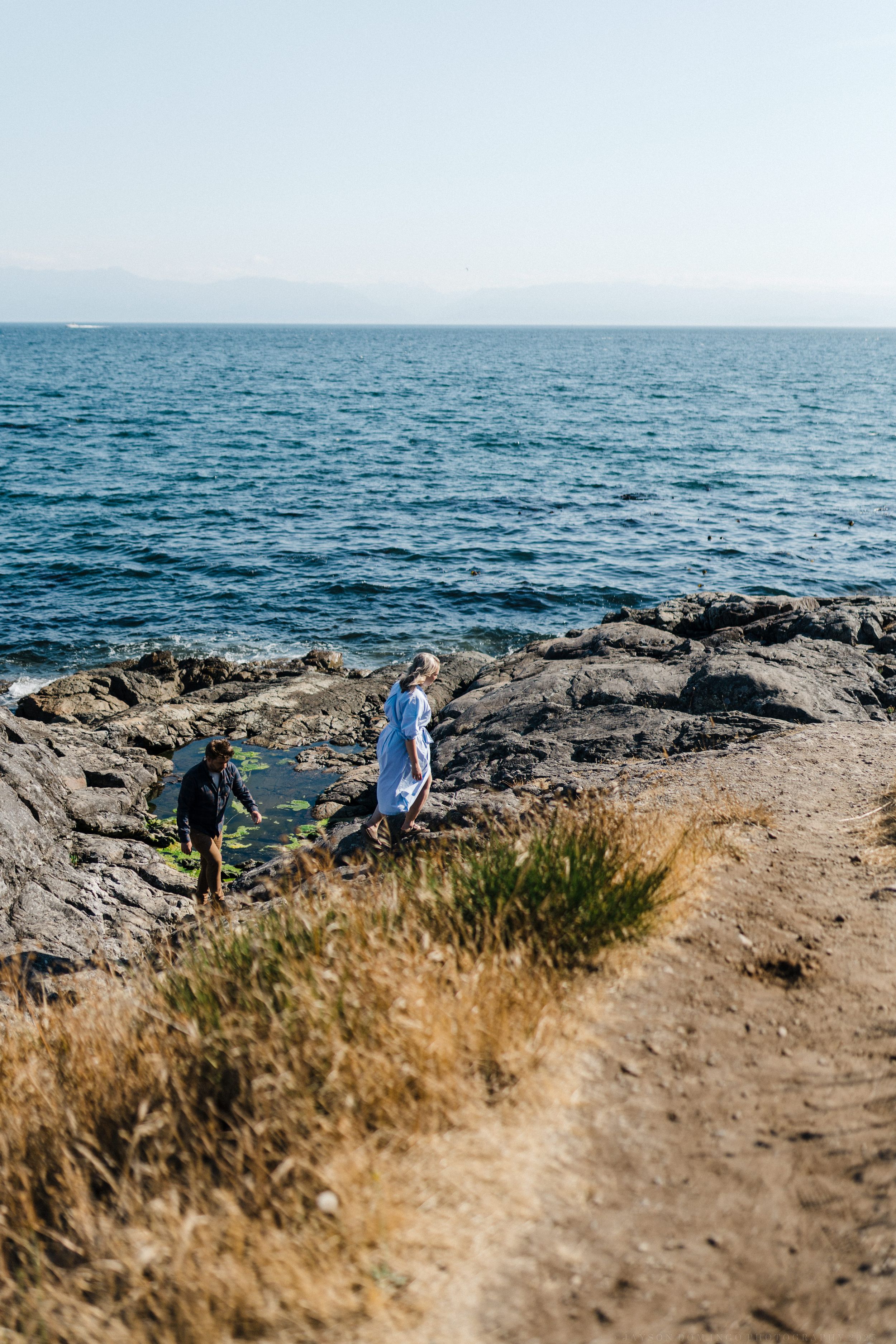 Two people exploring rocky coastal cliffs overlooking a vast blue ocean on a sunny day.