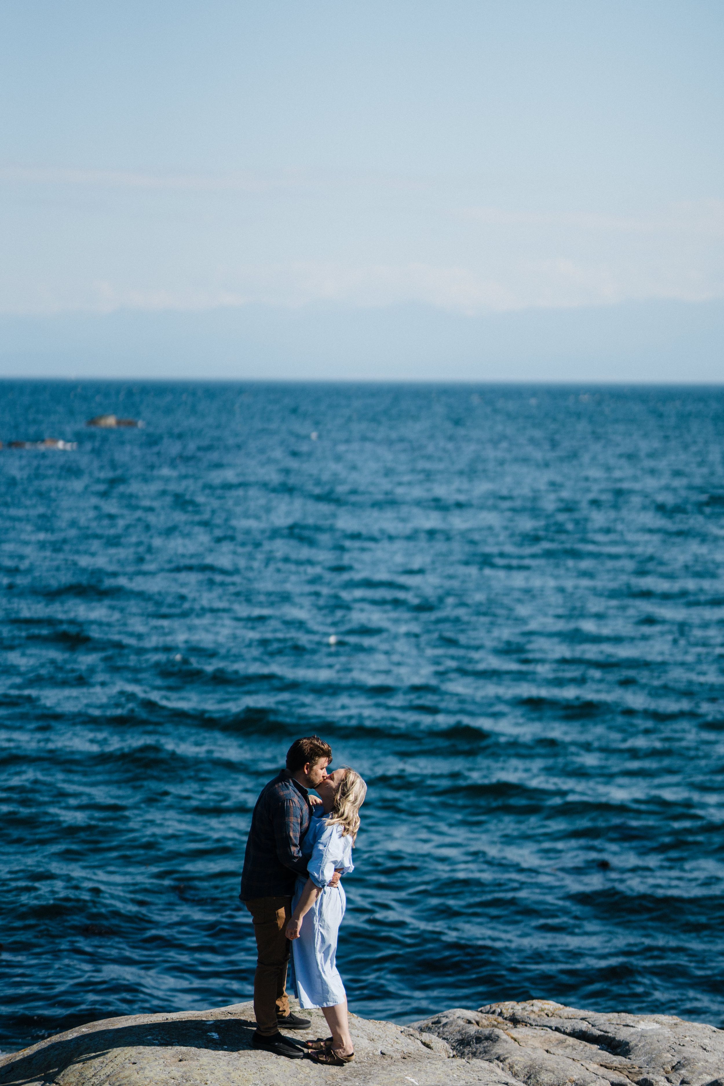 Two figures sit together on a rocky outcrop overlooking a vast blue ocean on a clear day.