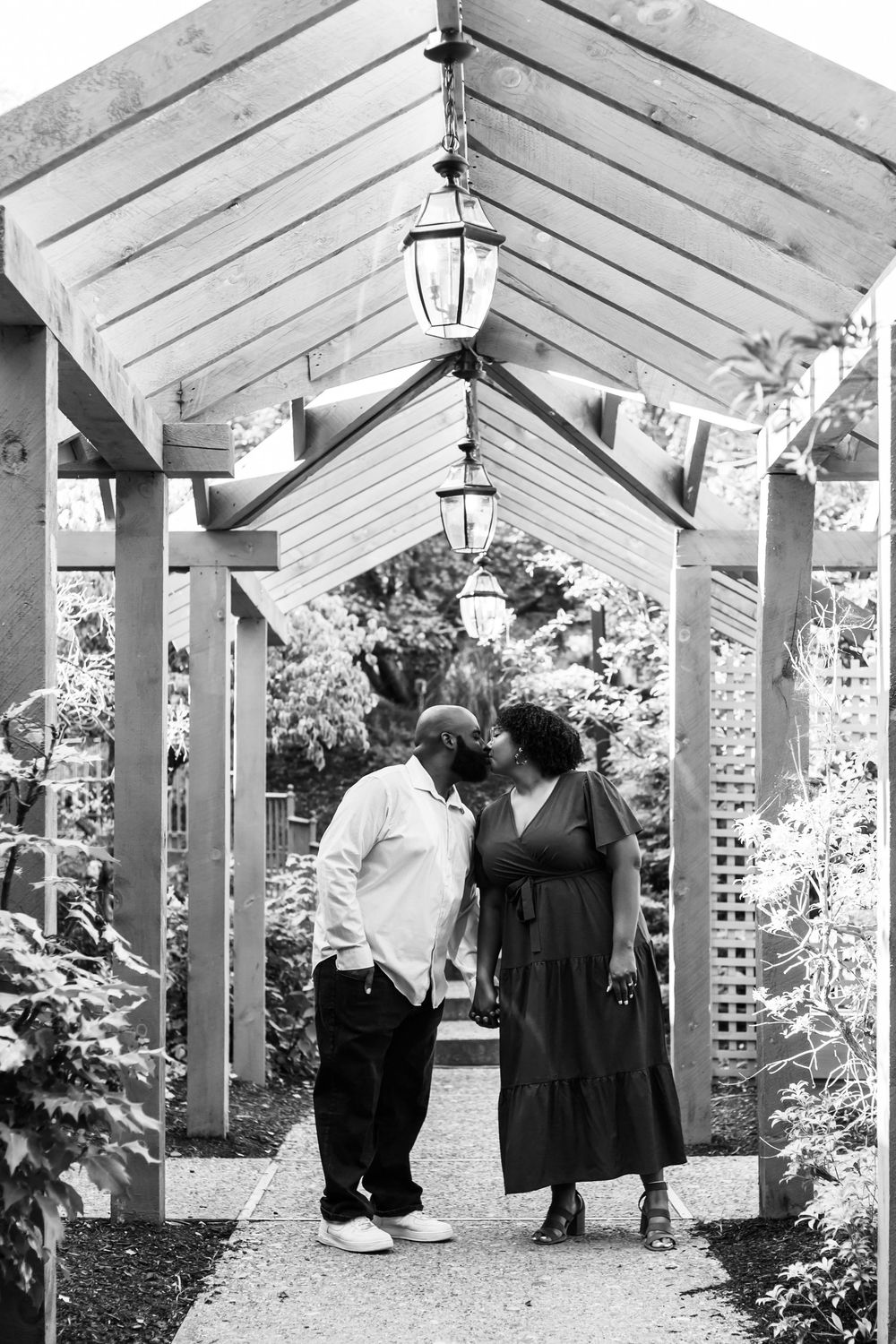 Couple in formal attire walking through an elegant white pergola garden path lined with hanging lights and greenery.