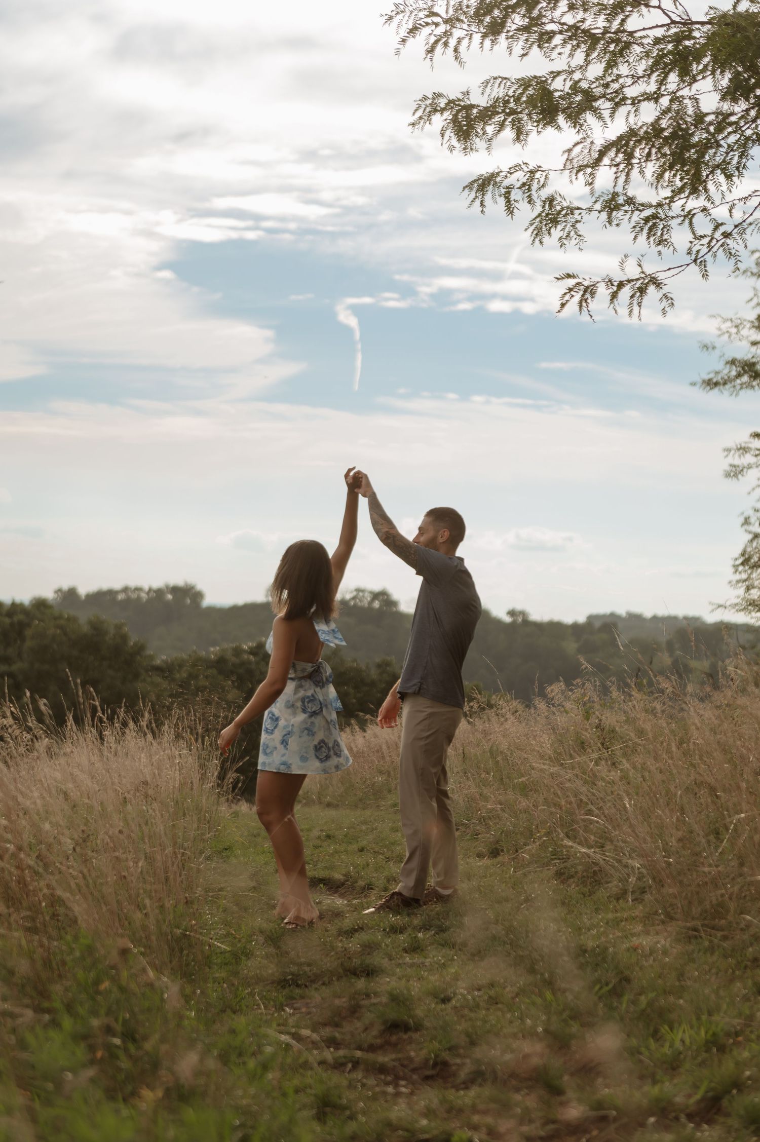 Couple dancing together in a scenic meadow field, holding hands raised, surrounded by tall grass and trees under a cloudy sky.