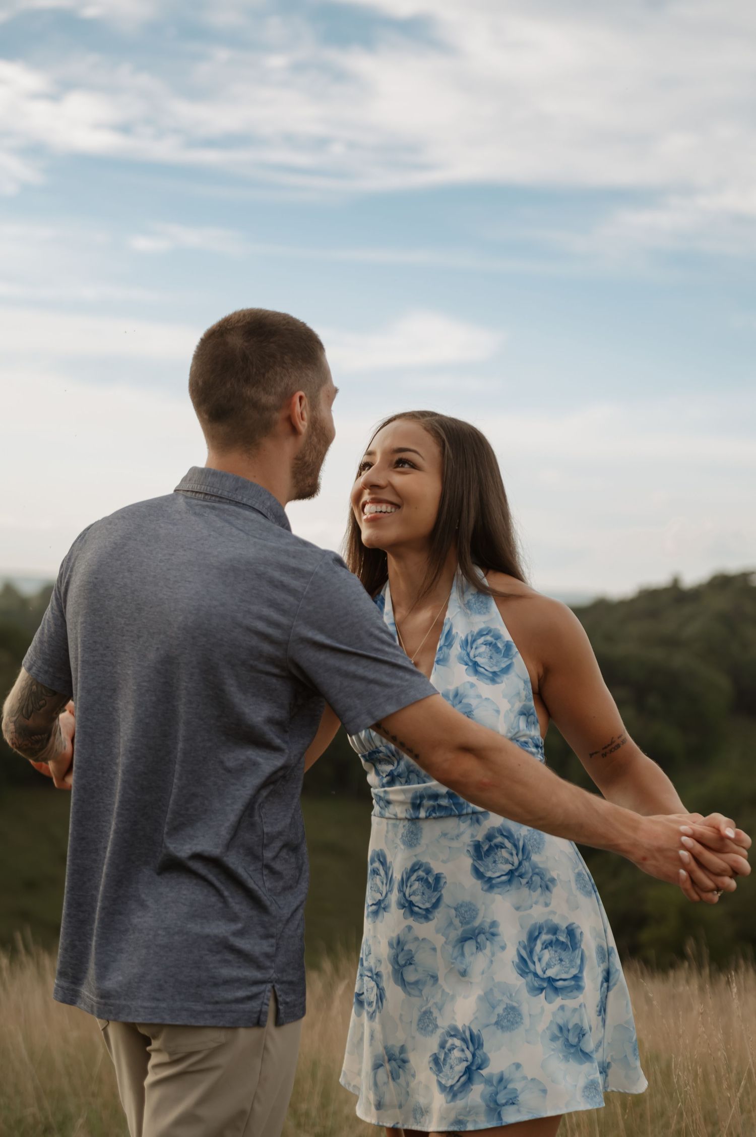 A smiling couple holds hands and gazes at each other in a grassy field, woman wearing a blue floral dress.