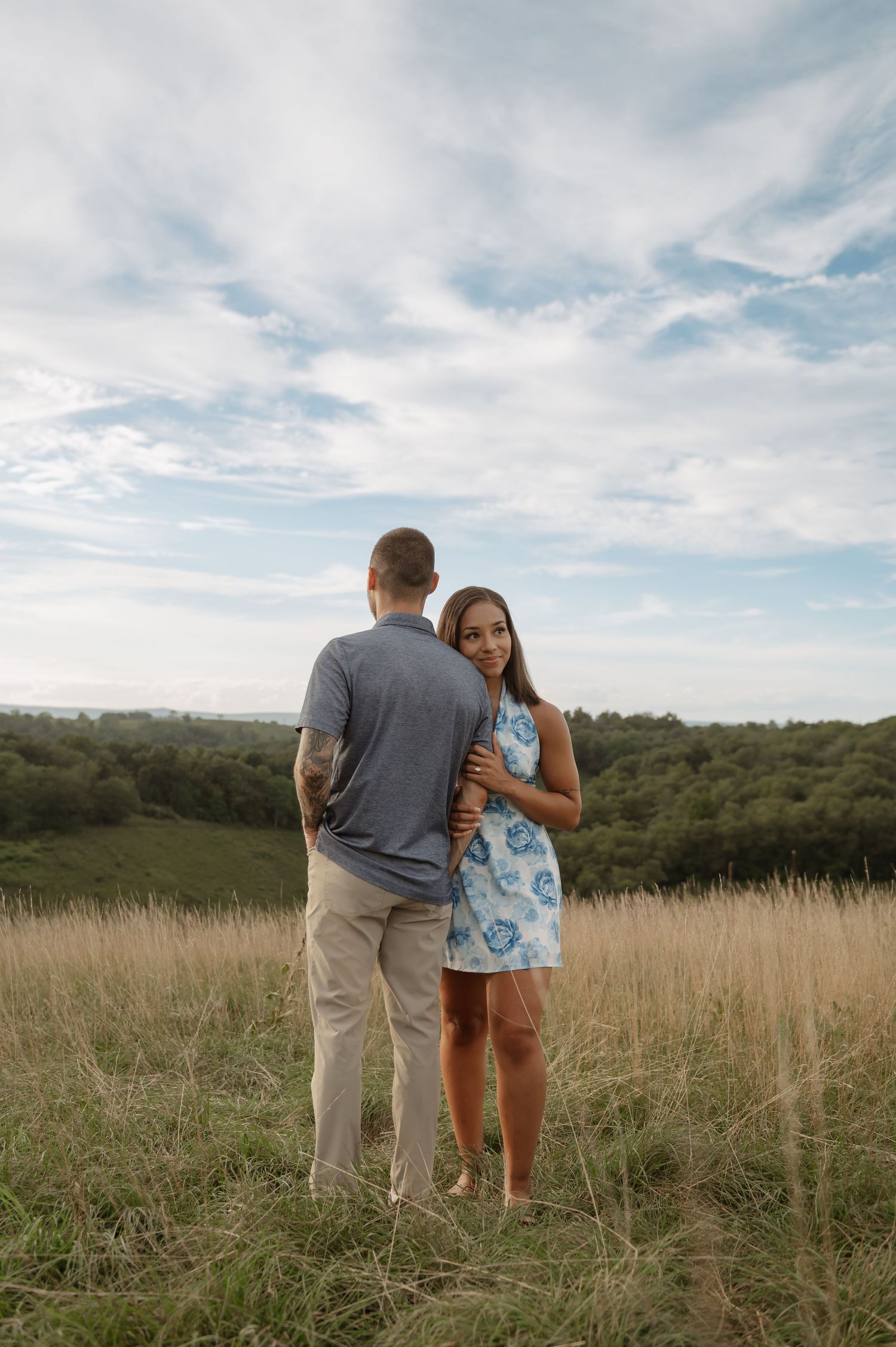 Couple embracing in a golden meadow, woman in blue floral dress smiling at camera, lush green hills and cloudy sky behind them.