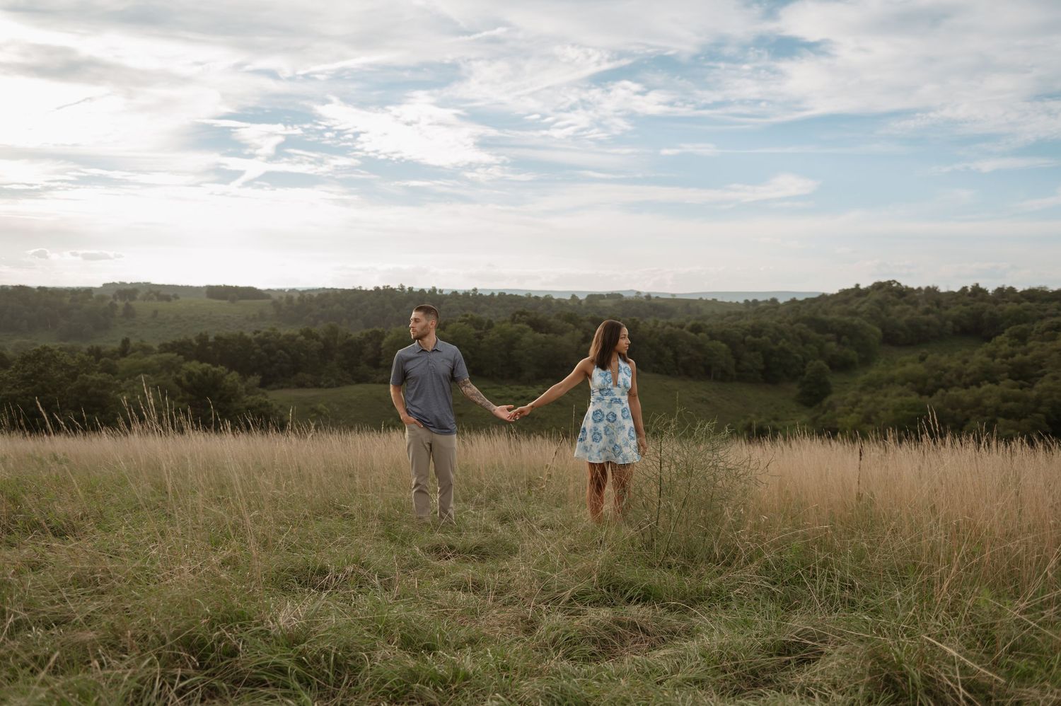 Couple holding hands in a golden grass field with lush green hills and cloudy sky backdrop.