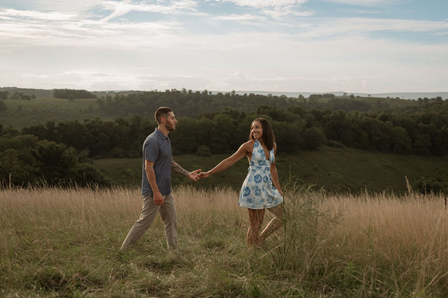 Couple holding hands walking through tall grass field with scenic green hills and cloudy sky in background.