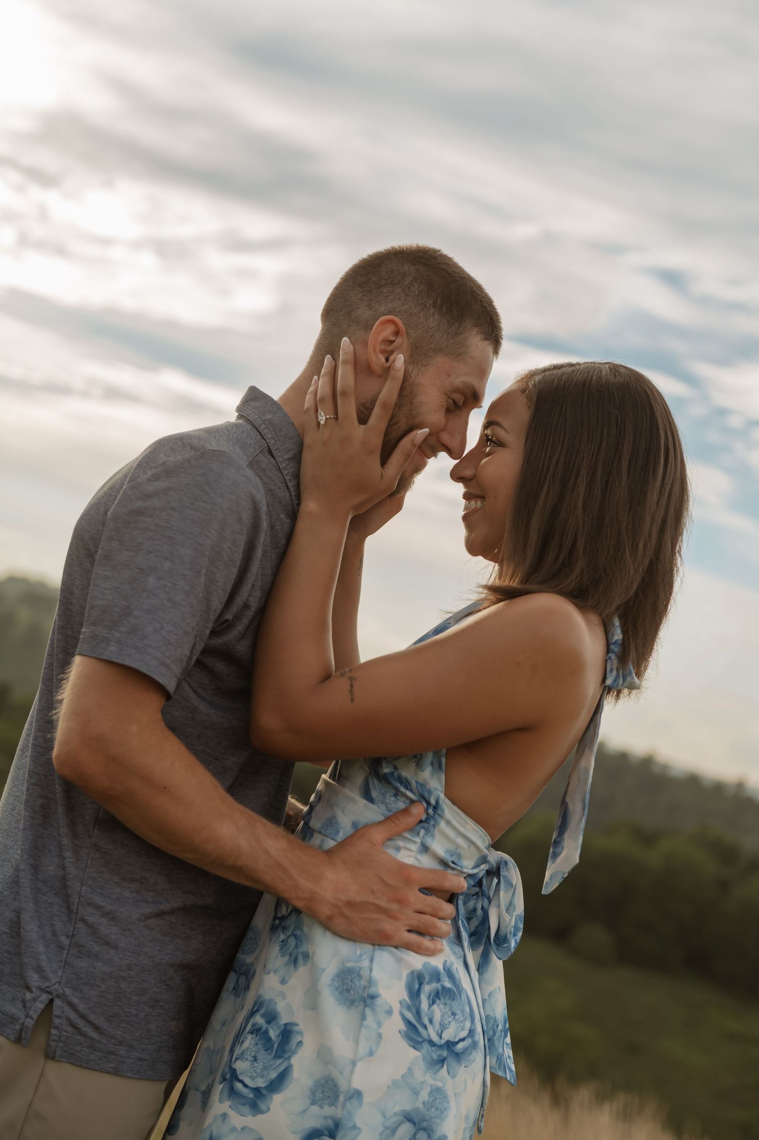 A couple shares a romantic moment outdoors, nearly kissing, with the woman in a floral dress and man in a gray shirt.