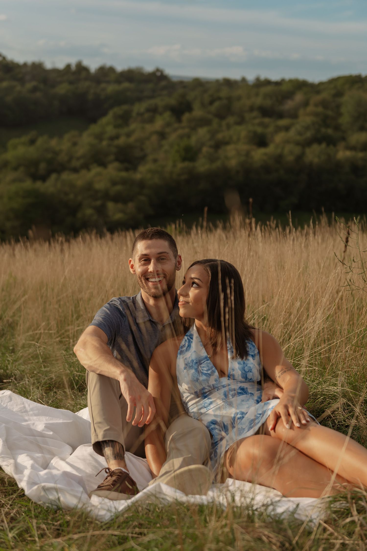 Happy couple sitting on a blanket in a sunny meadow with lush green hills in the background.