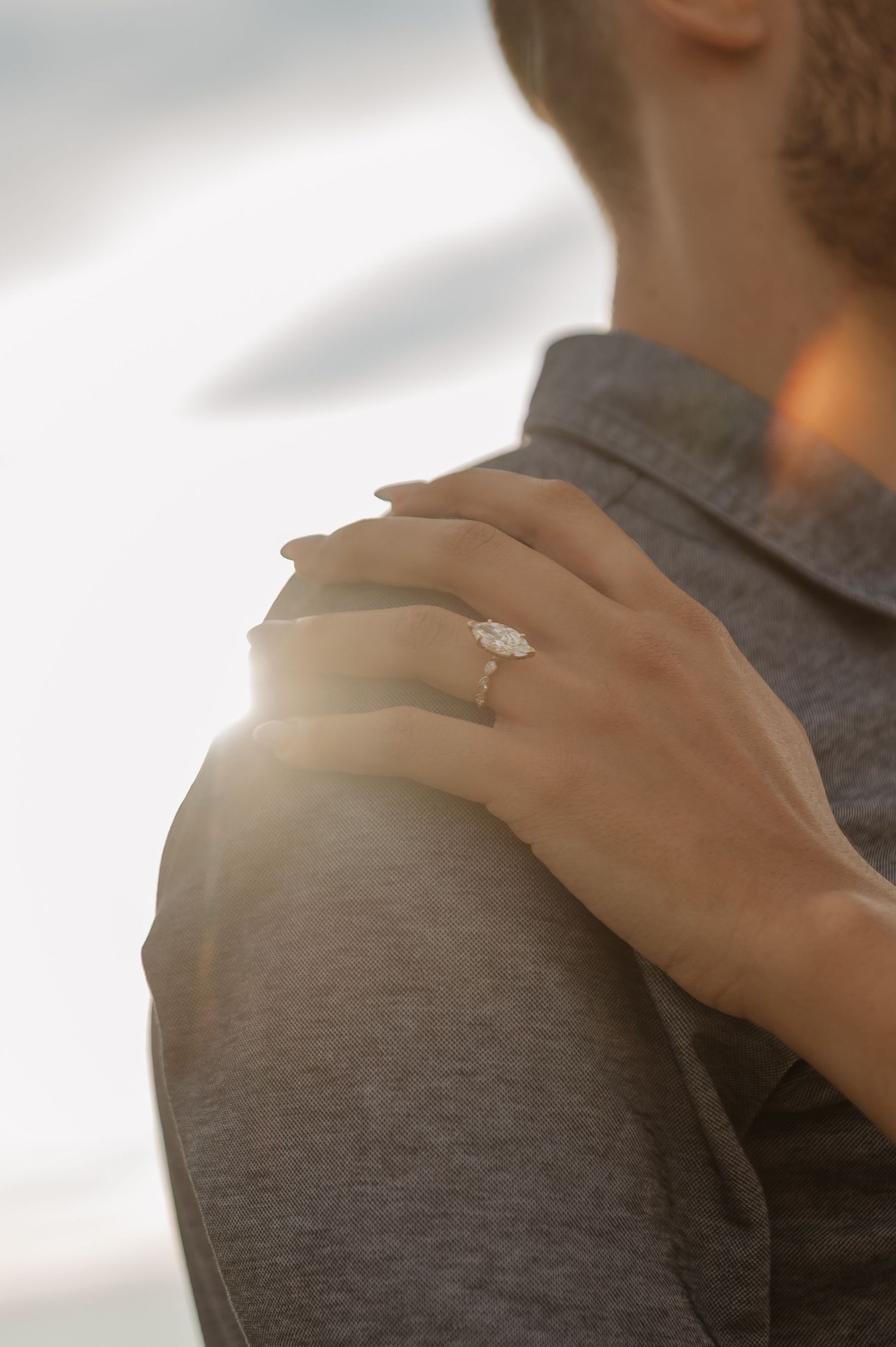 A woman rests her hand on a man's shoulder, showcasing a sparkling diamond engagement ring in warm sunset light.