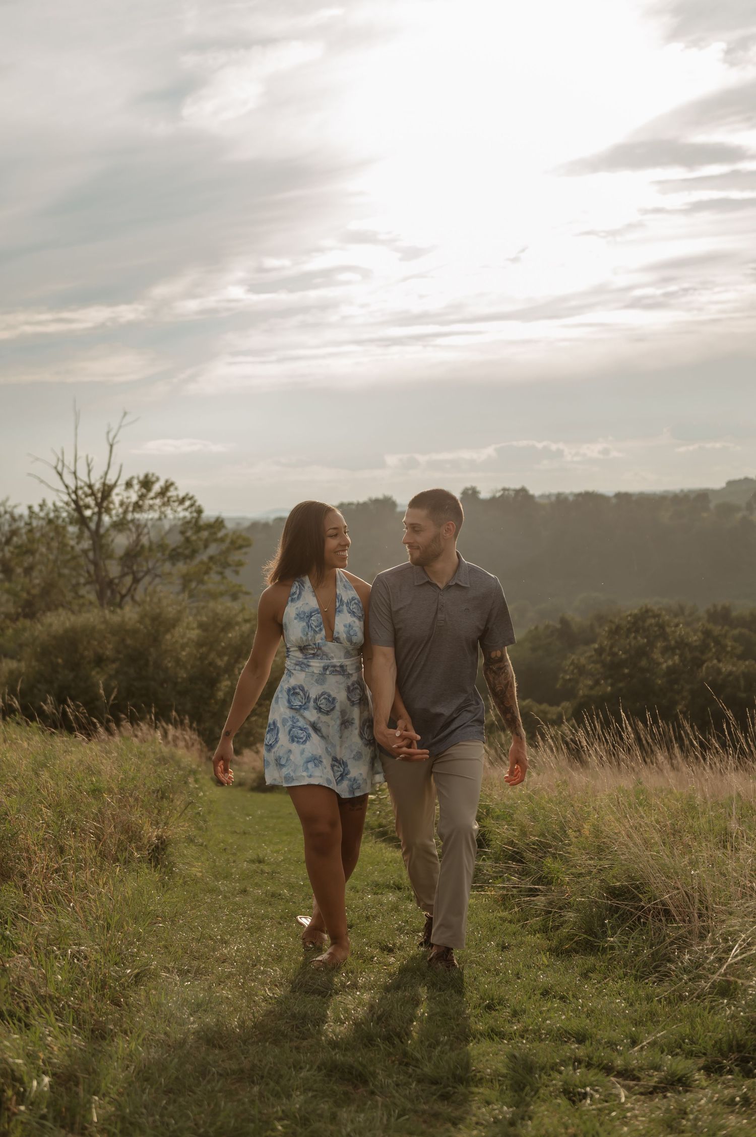 Couple holding hands walking through a scenic grassy hillside path at golden hour, woman in blue floral dress.