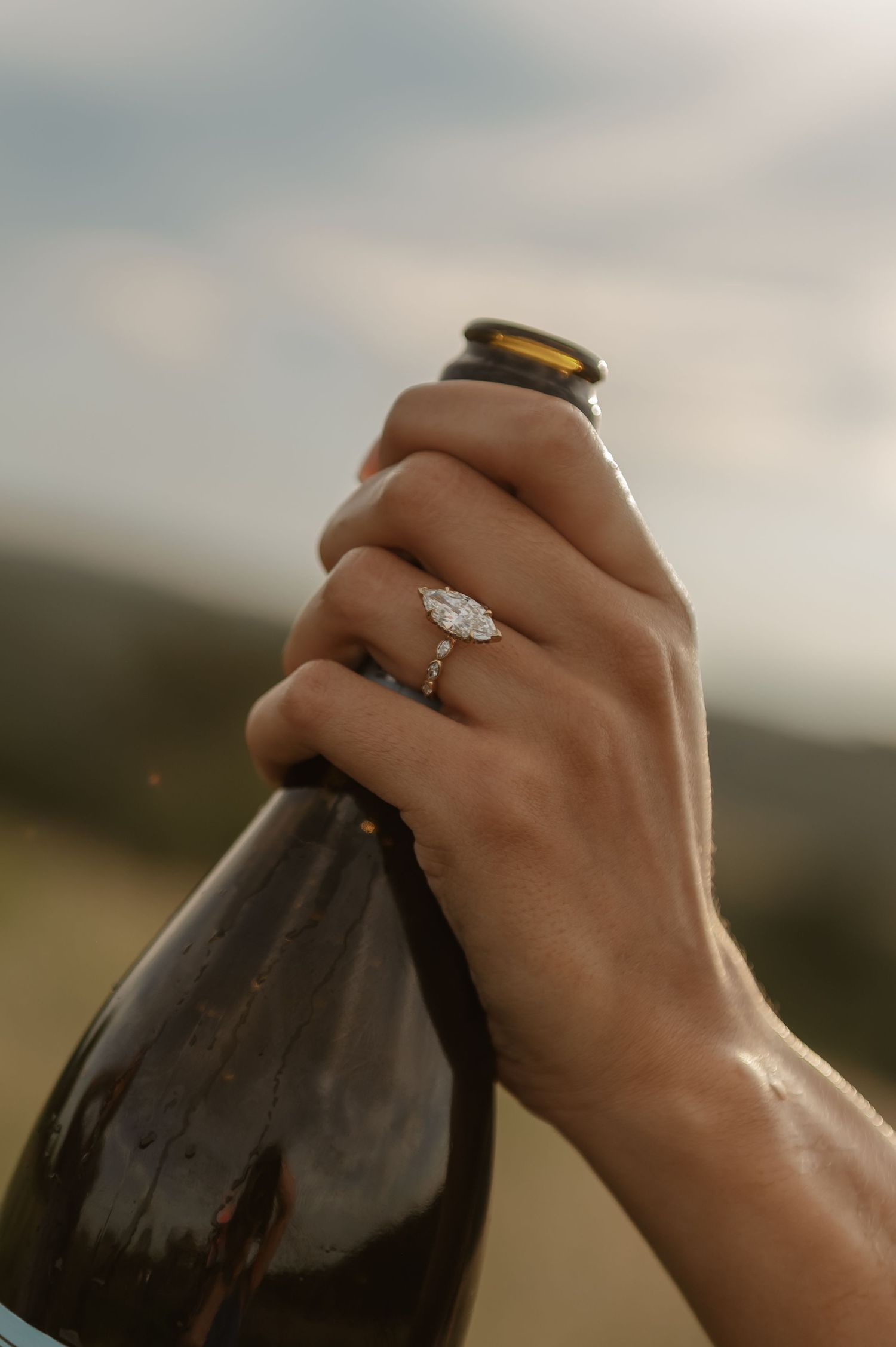 Close-up of hand holding dark wine bottle with elegant marquise diamond engagement ring in natural light.