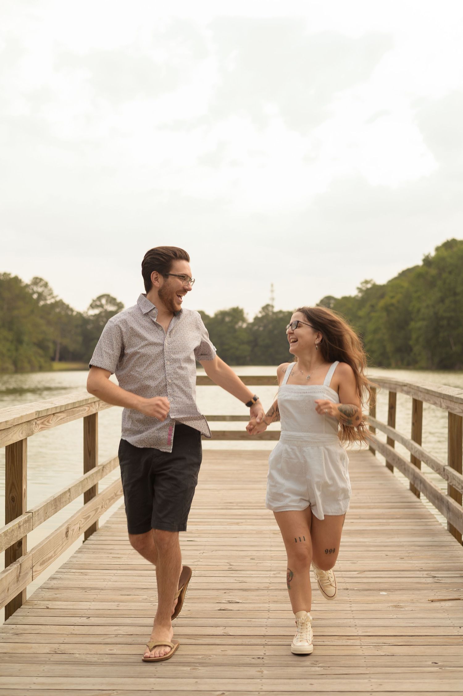 A couple playfully chases each other on a wooden boardwalk near a lake during sunset.