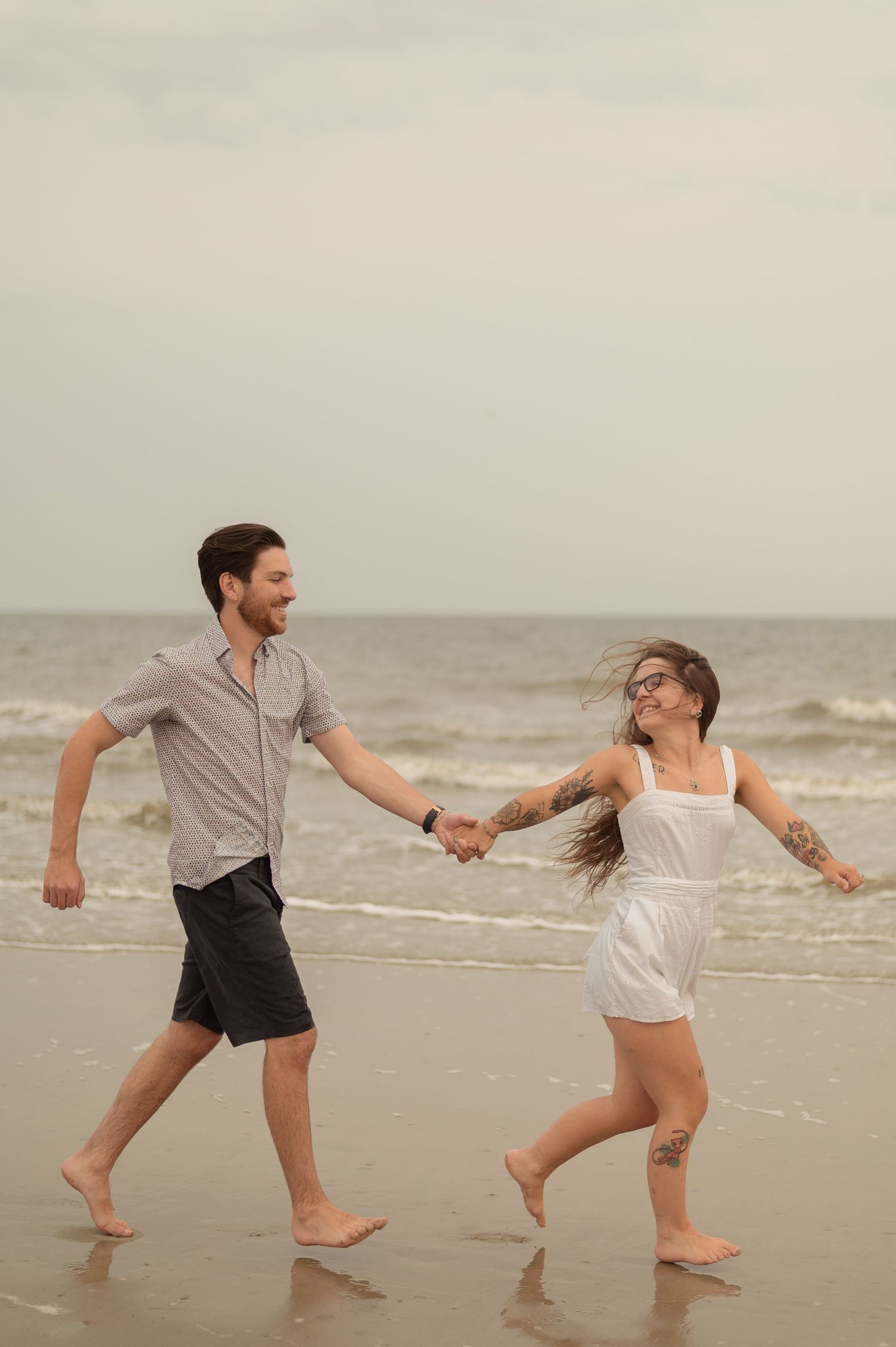 A couple holds hands while joyfully running barefoot along a misty beach shore.
