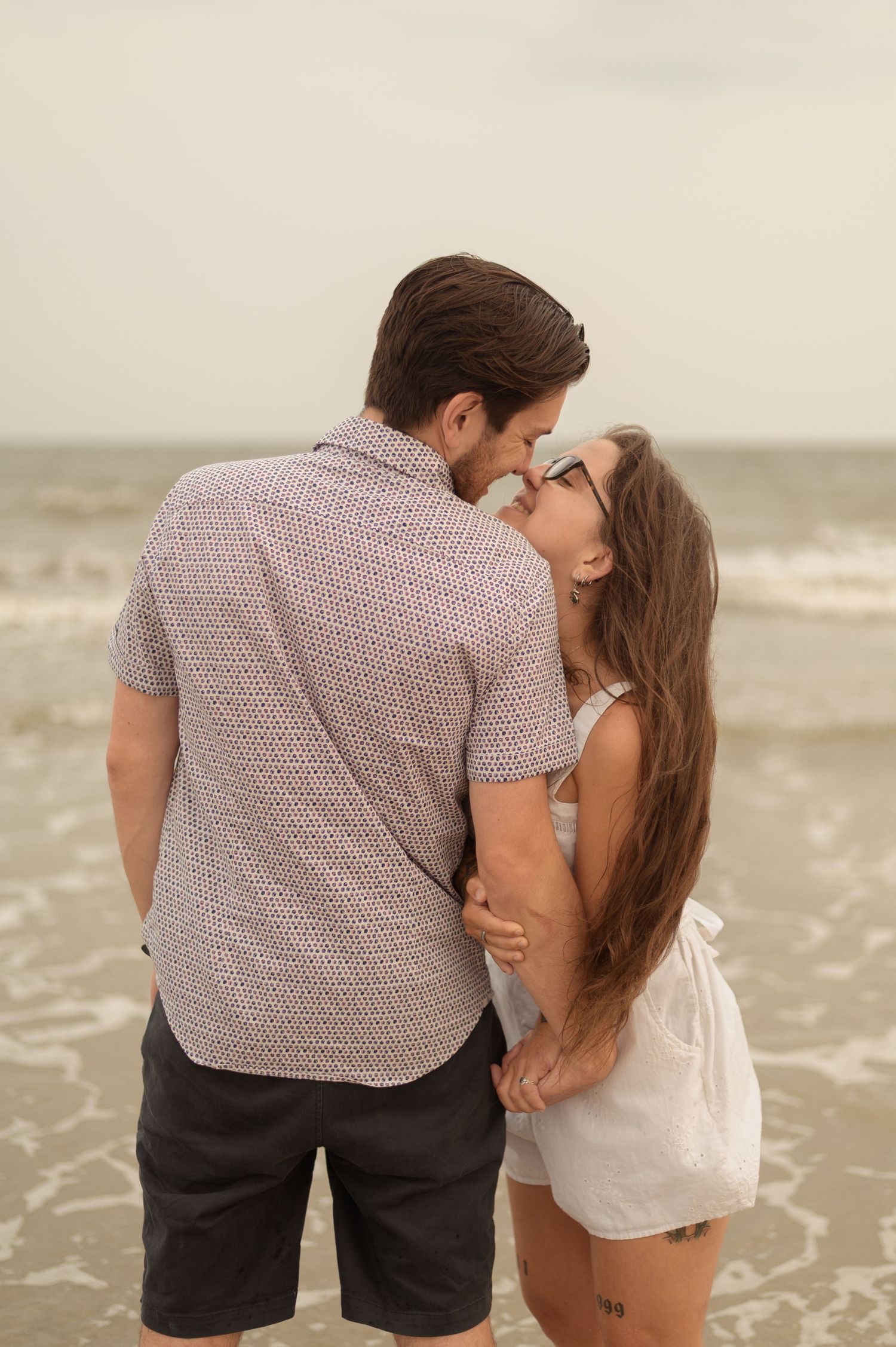 A romantic couple shares a tender kiss on a sandy beach with ocean waves in the background.