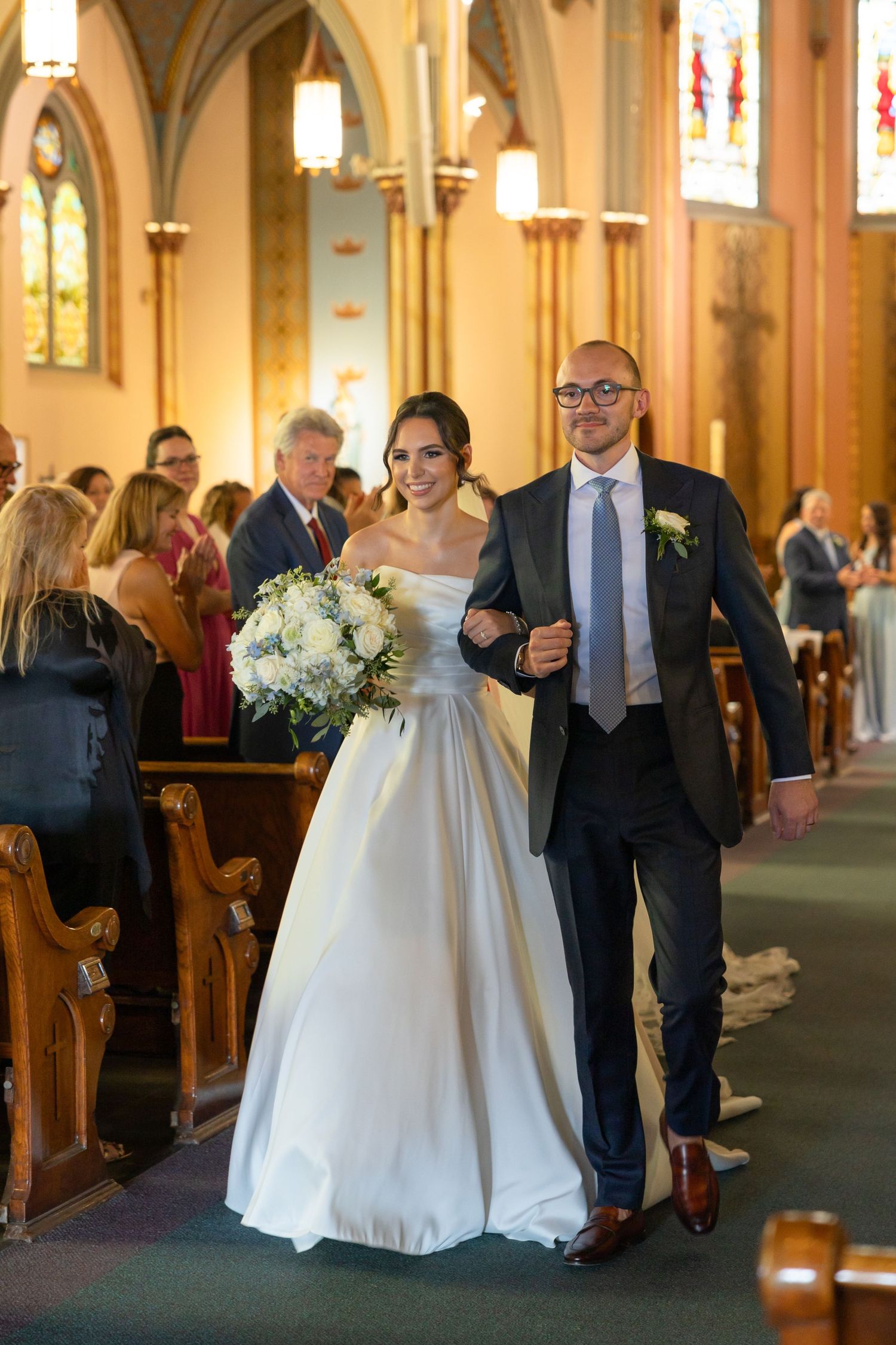 Traditional wedding ceremony procession walks down the church aisle decorated with beautiful stained glass windows. Rachel Martinez