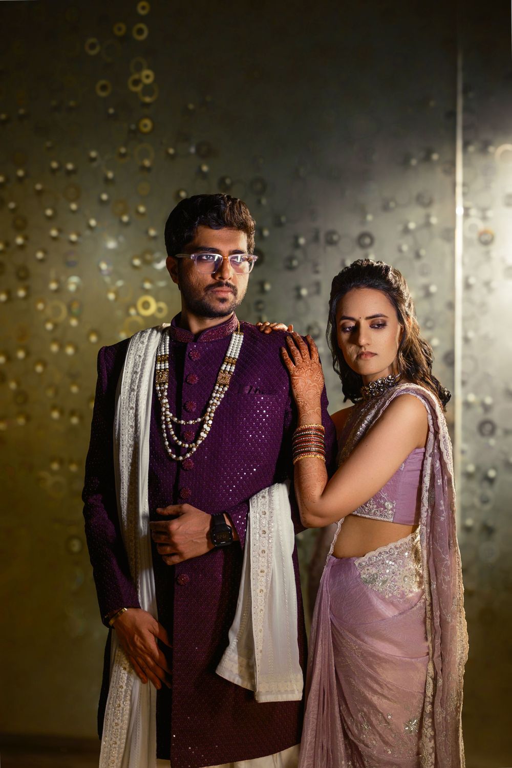 Couple in traditional Indian wedding attire pose together against a sparkly backdrop wearing purple and pink outfits.