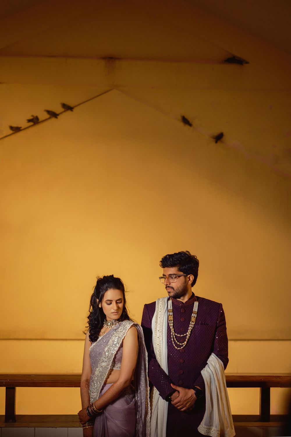 A couple in traditional Indian attire pose dramatically against a warm sunset wall with birds perched on wires above.