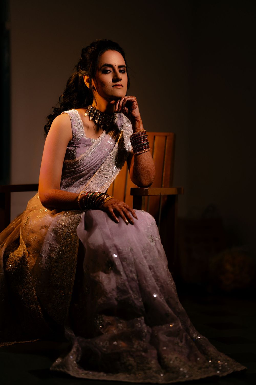 Bride in elegant silver saree poses thoughtfully on wooden chair in dramatic lighting.