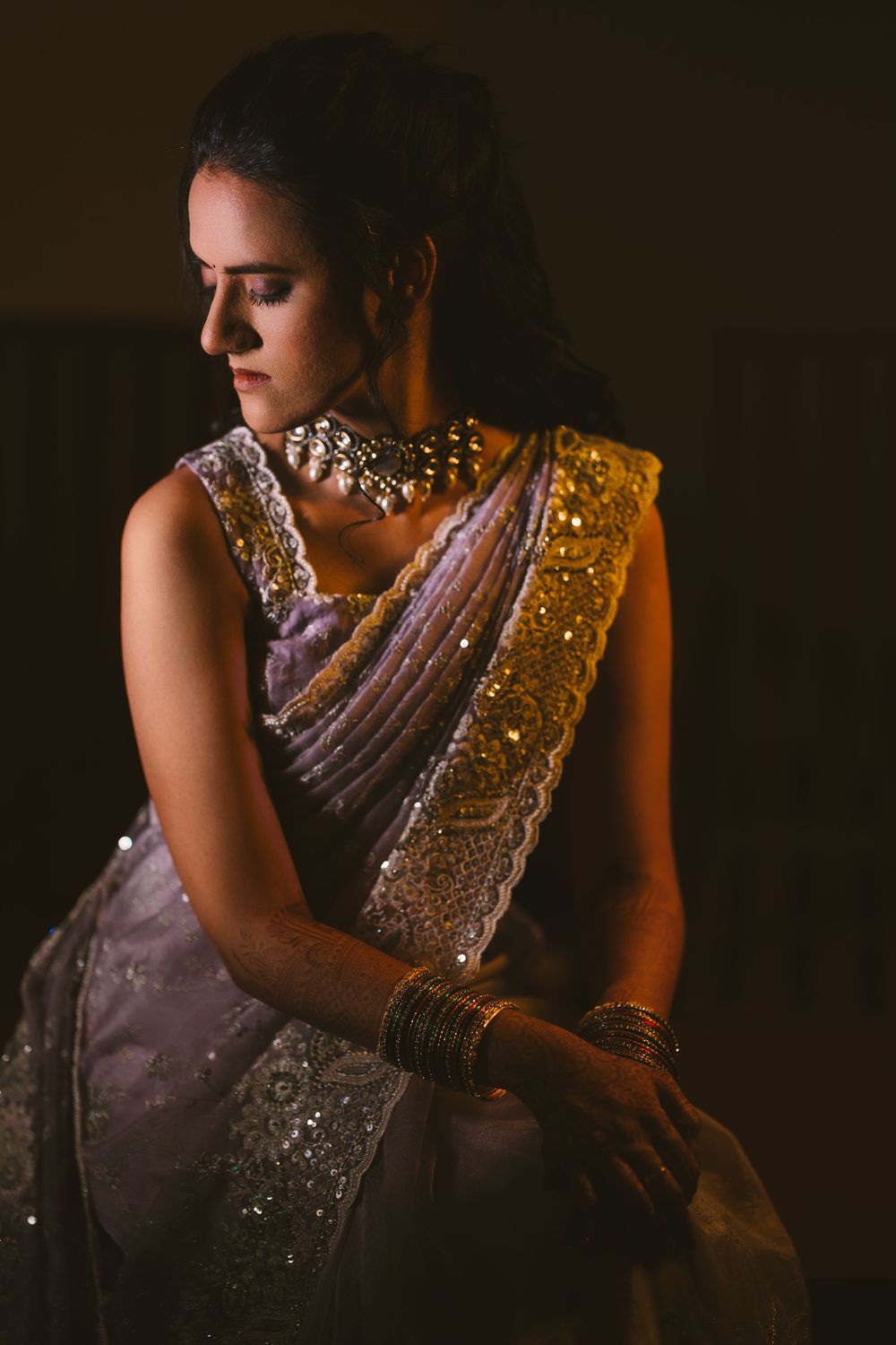 A dramatic portrait showing intricate details of a sparkly purple and gold sari with statement jewelry in moody lighting.
