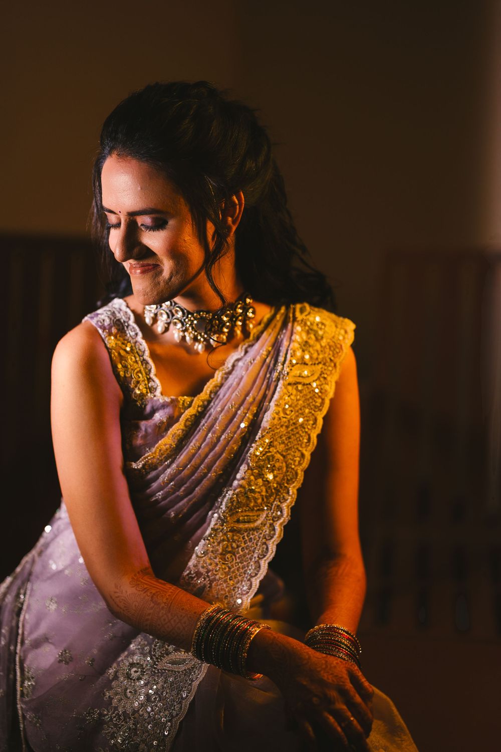 Person in lavender sari with intricate embellishments and jewelry poses in dramatic lighting.