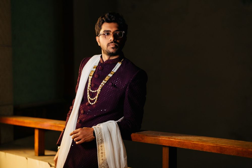 Traditional Indian groom poses in purple sherwani with white dhoti and beaded necklace in dramatic lighting.