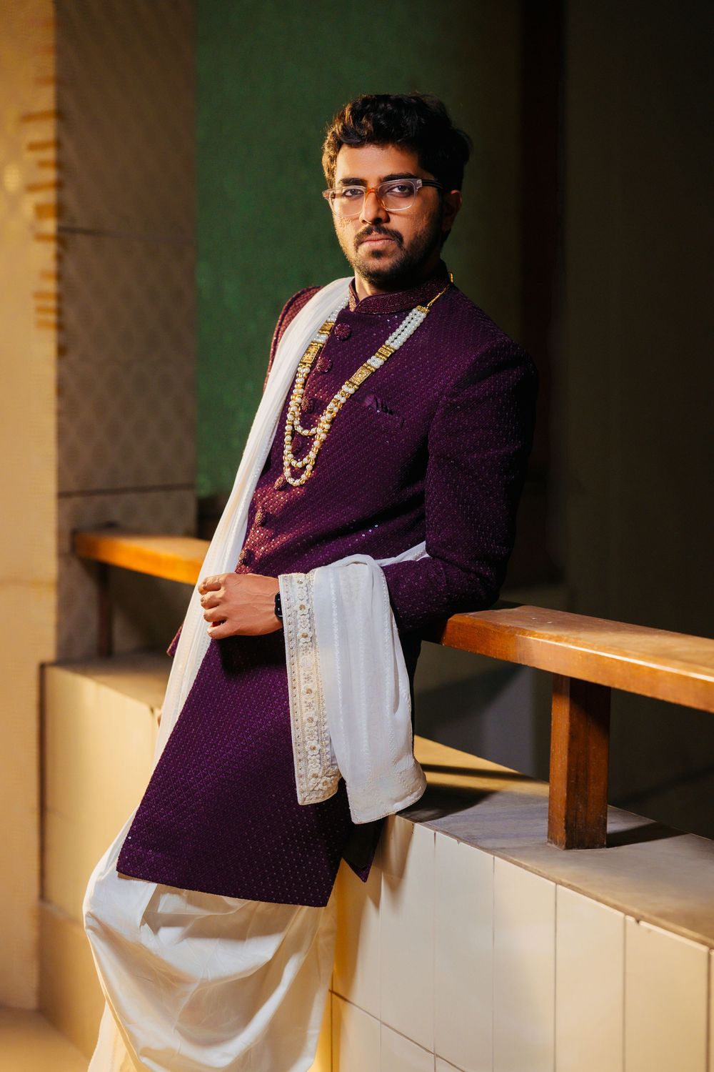 Elegant wedding attire featuring purple sherwani, white dupatta, and traditional Indian jewelry against green wall.
