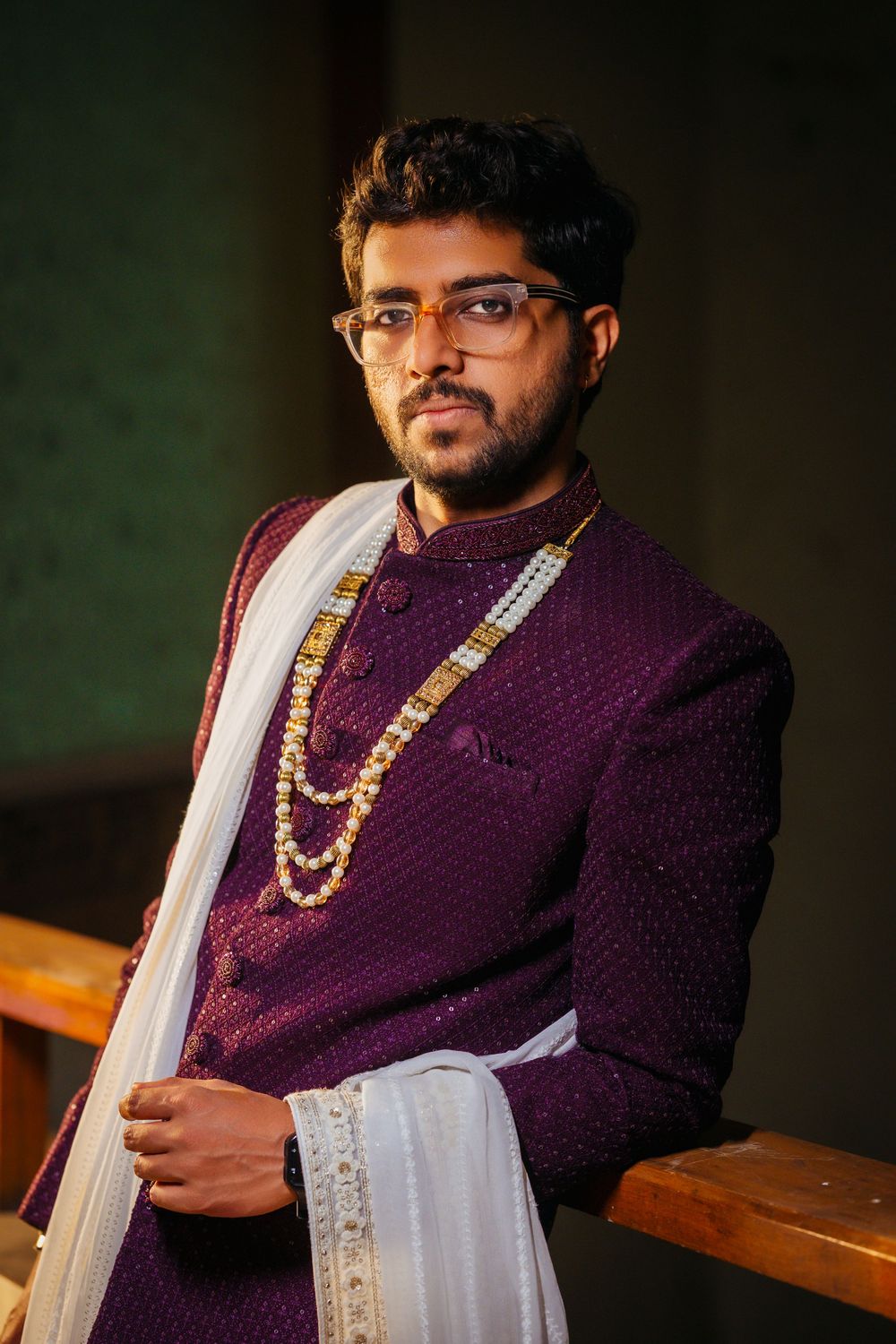 Traditional Indian groom in a deep purple sherwani and white dupatta with gold necklace against dark background.