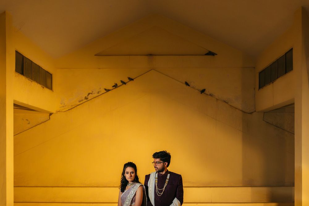 Couple stands against a yellow wall in a minimalist setting with dramatic lighting and shadows.