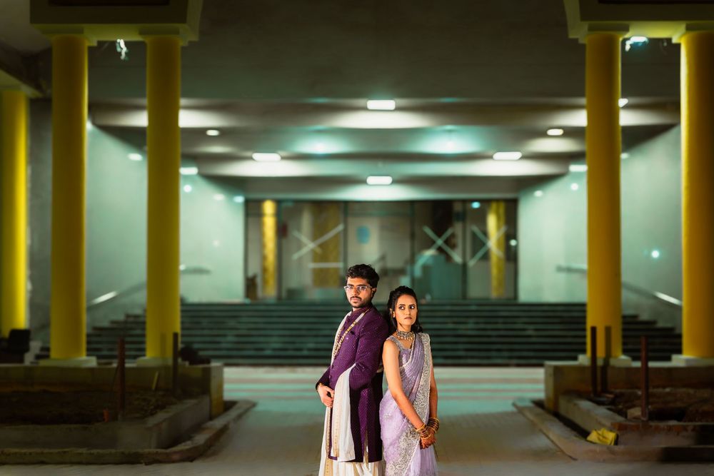 A couple in coordinated ethnic outfits stands in an illuminated indoor venue with yellow pillars and seating.