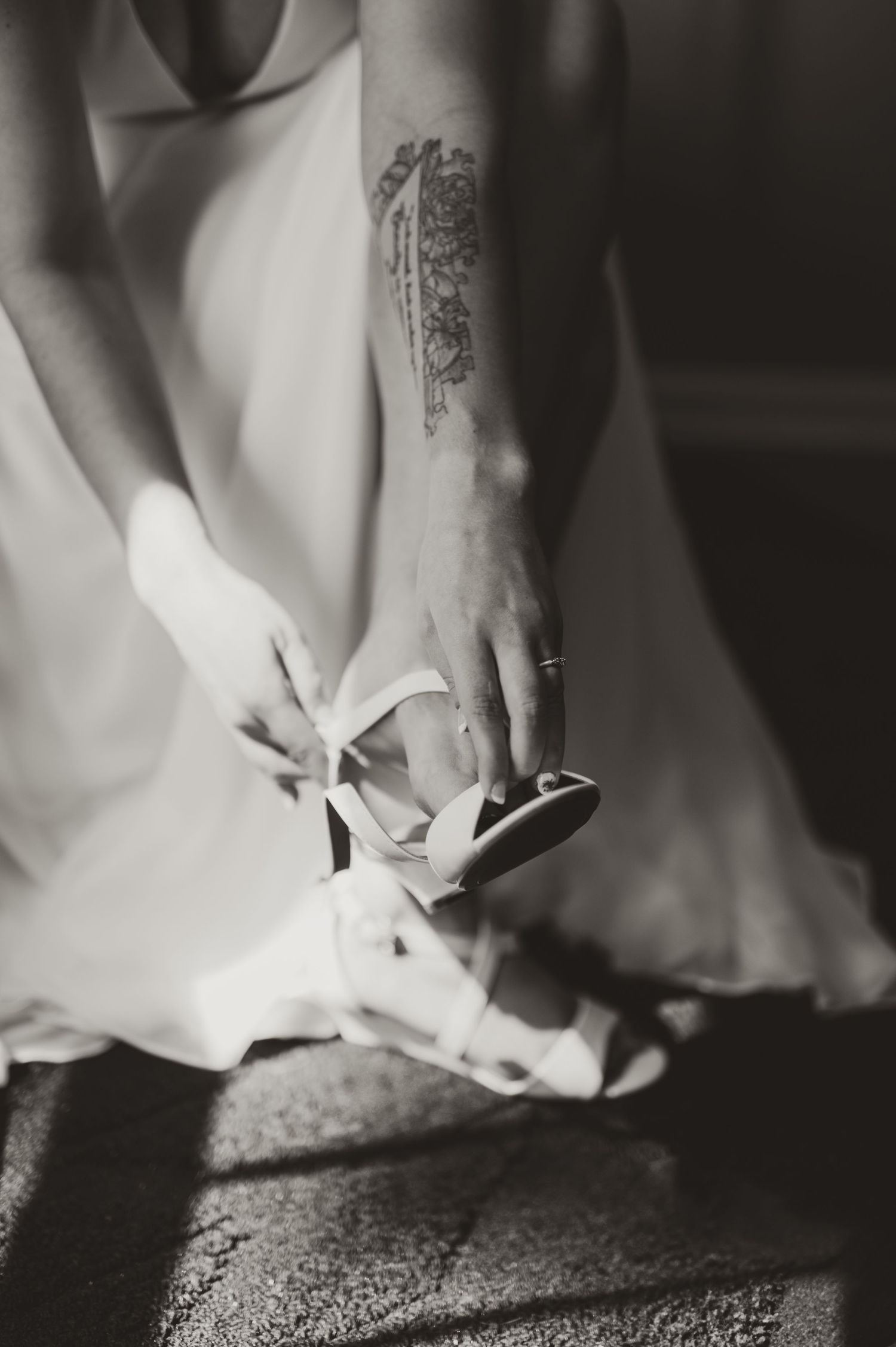 Bride with forearm tattoo fastening strappy heels in black and white wedding photo.