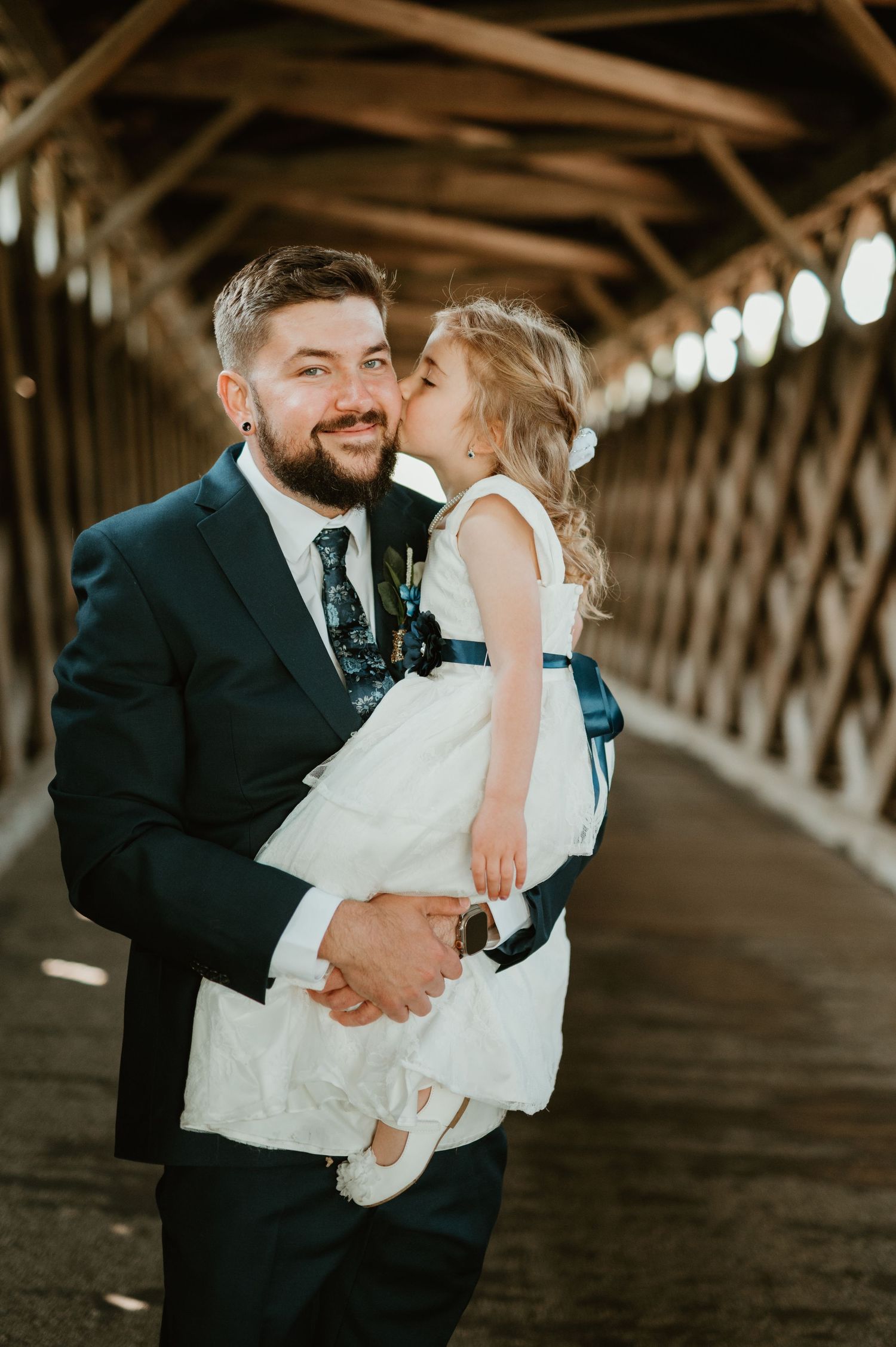 A bearded groom in a navy suit holds a young girl in a white dress who kisses his cheek inside a covered wooden bridge.