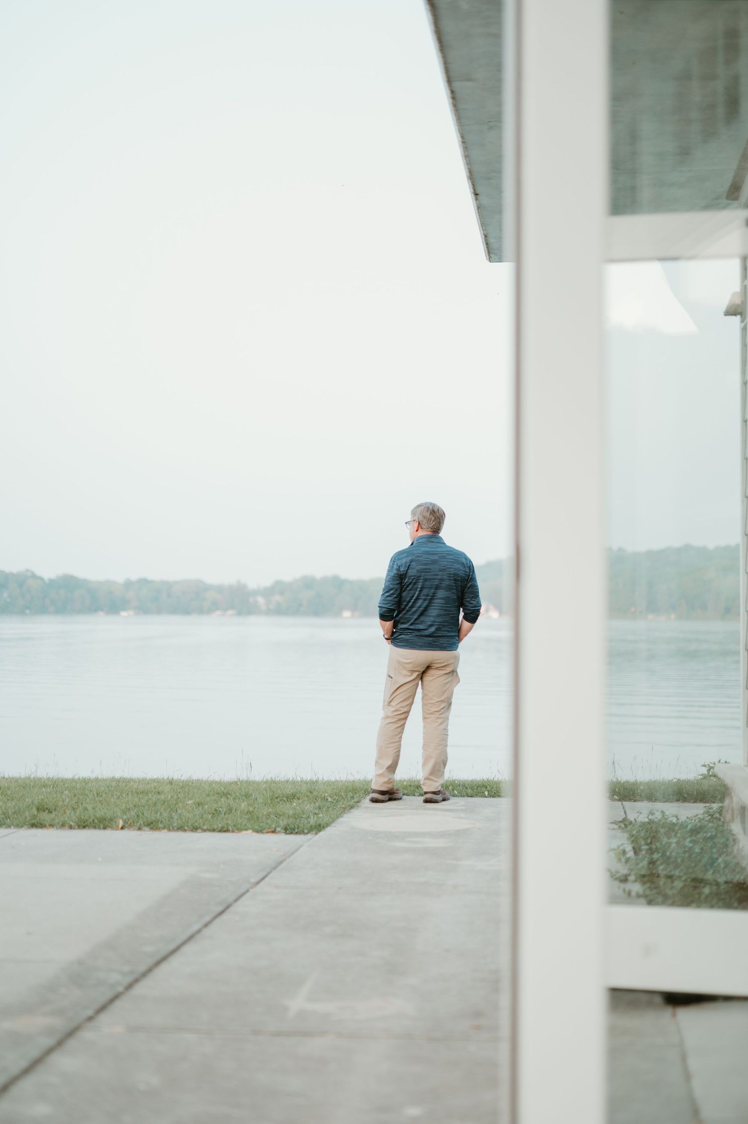 Elderly man in blue jacket and khaki pants stands on patio gazing at calm lake surrounded by lush trees in Hartford WIsconsin