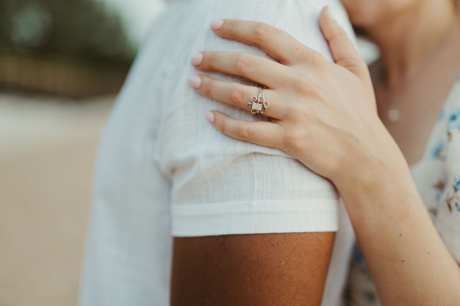Close-up of a woman's hand with a diamond engagement ring resting on a man's shoulder.