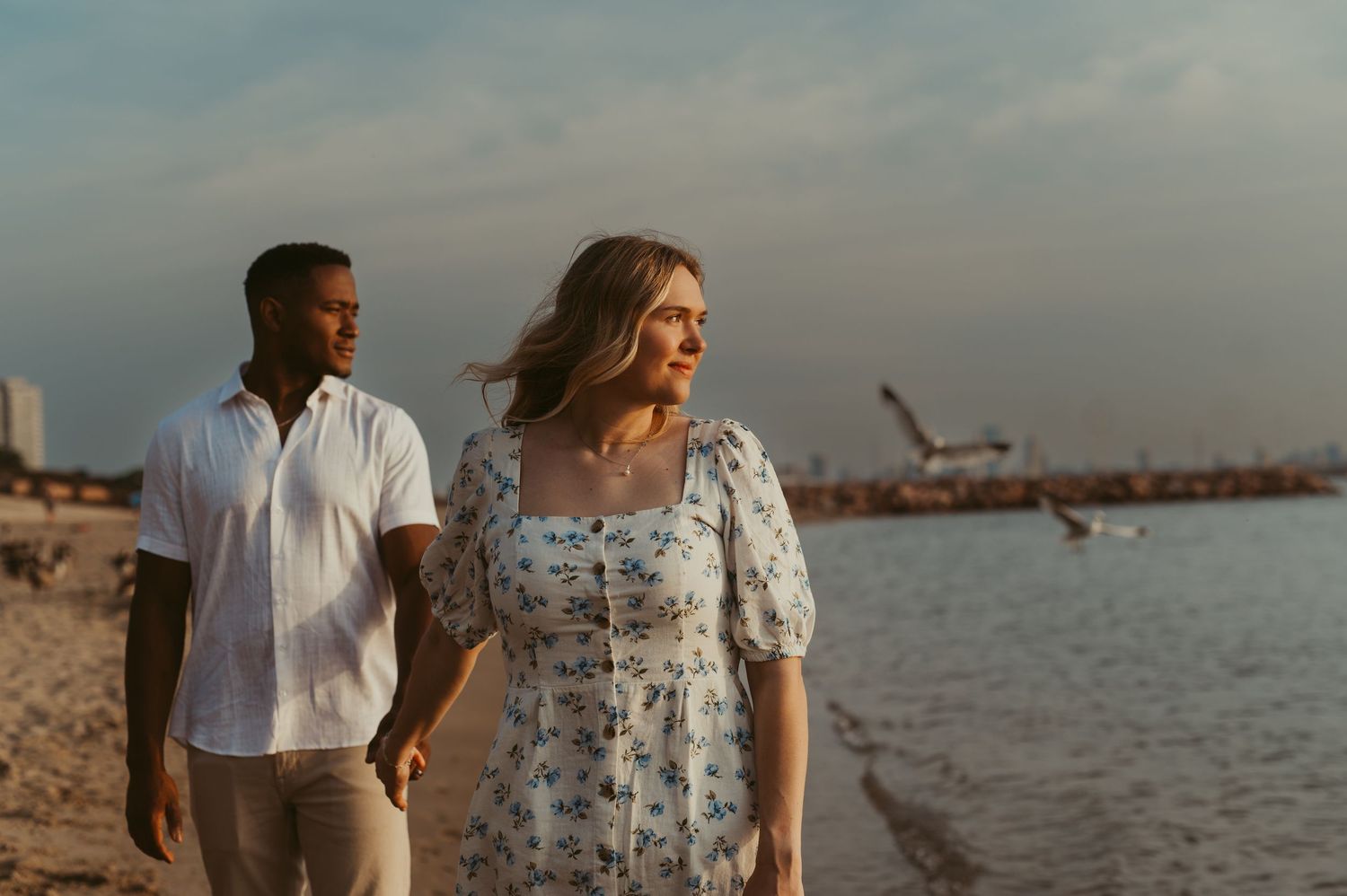 Couple holding hands walking on beach at sunset, woman in floral dress and man in white shirt.