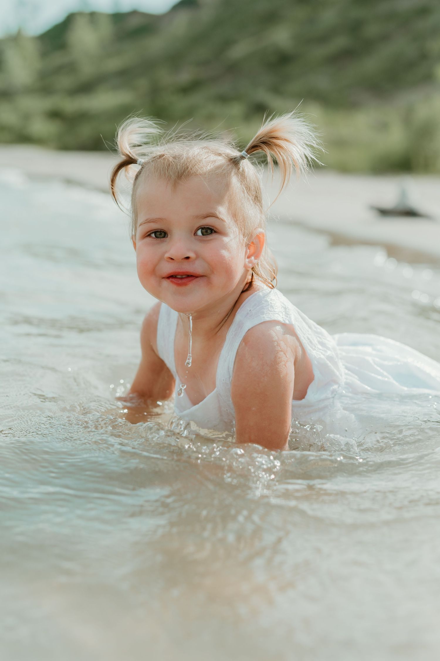 A smiling toddler girl with pigtails plays in shallow ocean waves wearing a white dress on a sandy beach.