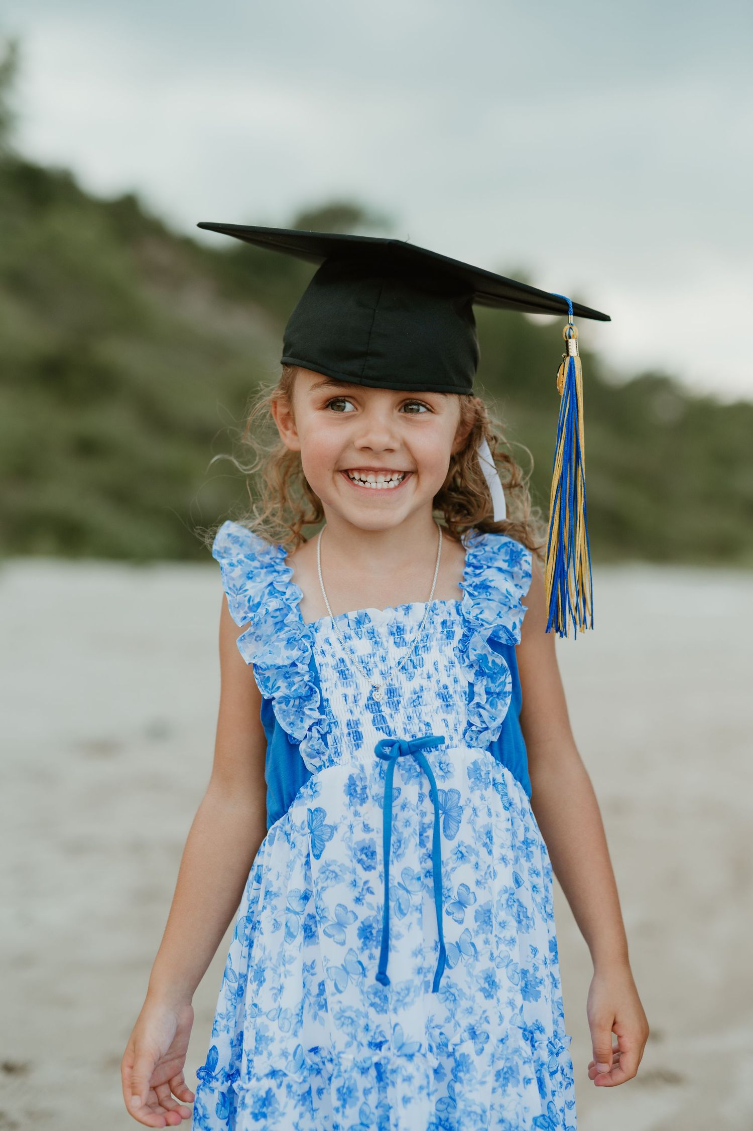 Young girl smiling in blue floral dress and graduation cap on sandy beach.