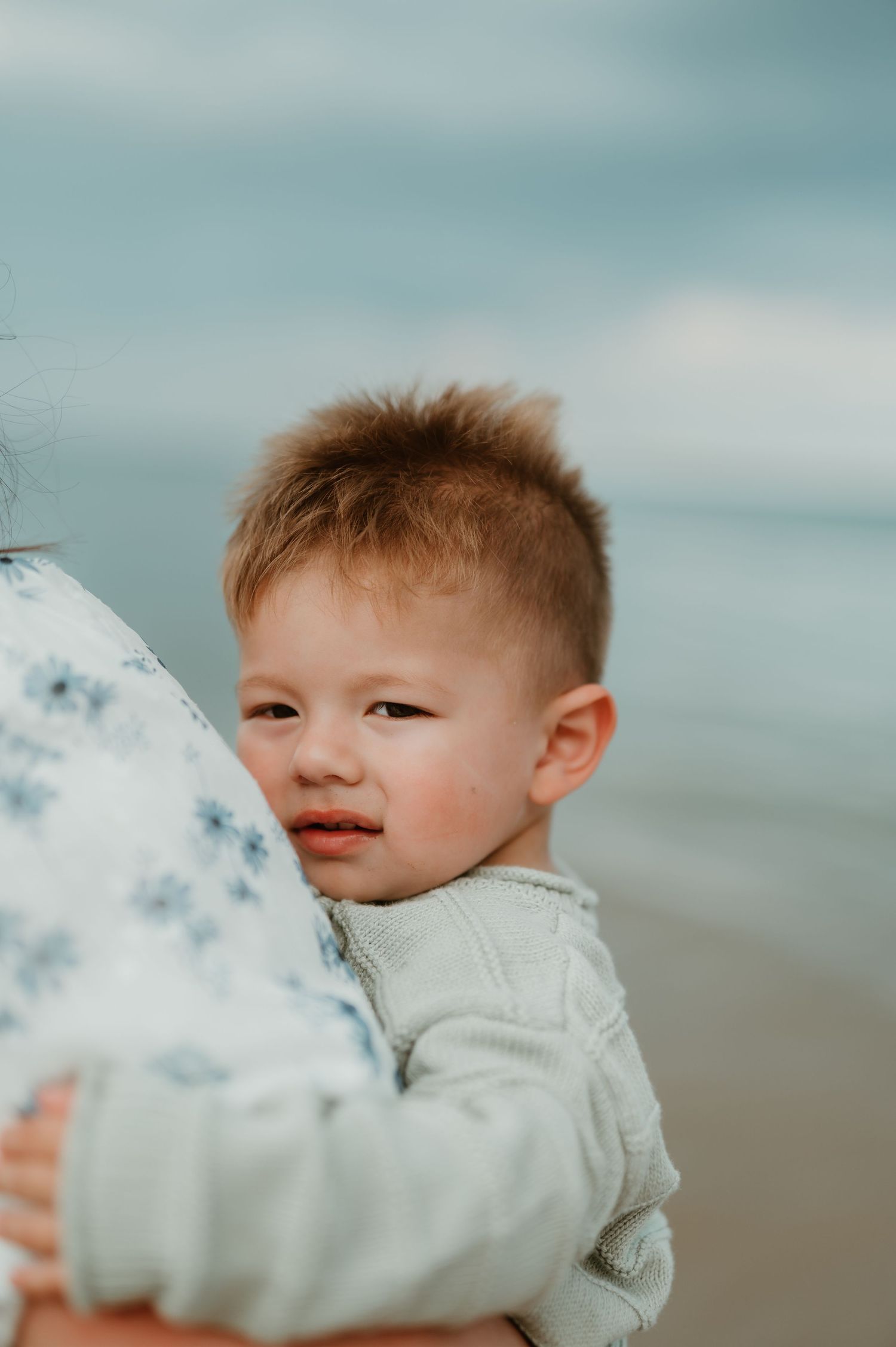 A young toddler boy with short hair clings to an adult at a beach, wearing a light sweater on a cloudy day.