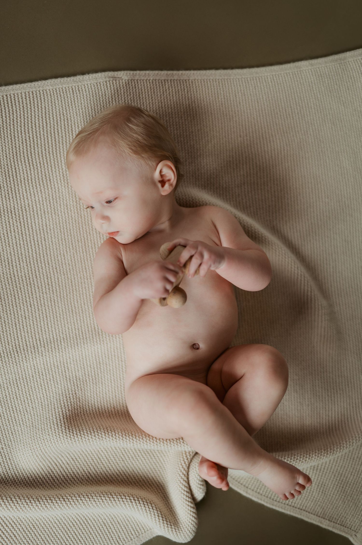 A peaceful newborn baby lying on a soft cream knit blanket, holding a small wooden toy.