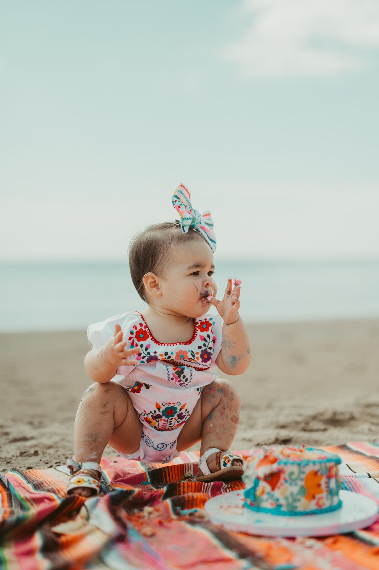 Baby girl in floral romper and bow headband sitting on beach blanket with birthday cake at her first birthday celebration.