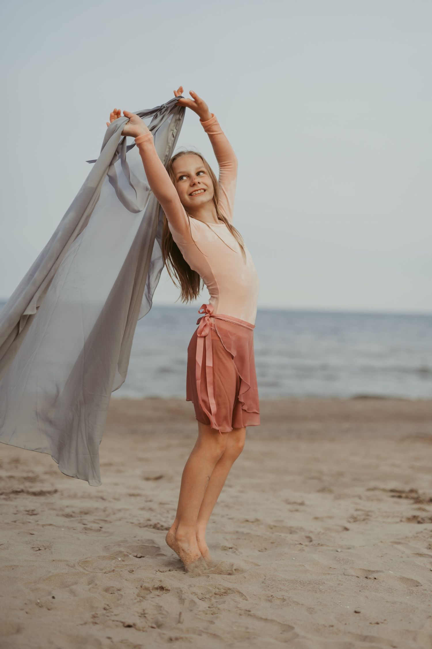 Young girl dancing on sandy beach holding flowing gray fabric, wearing pink top and skirt, with ocean in background.
