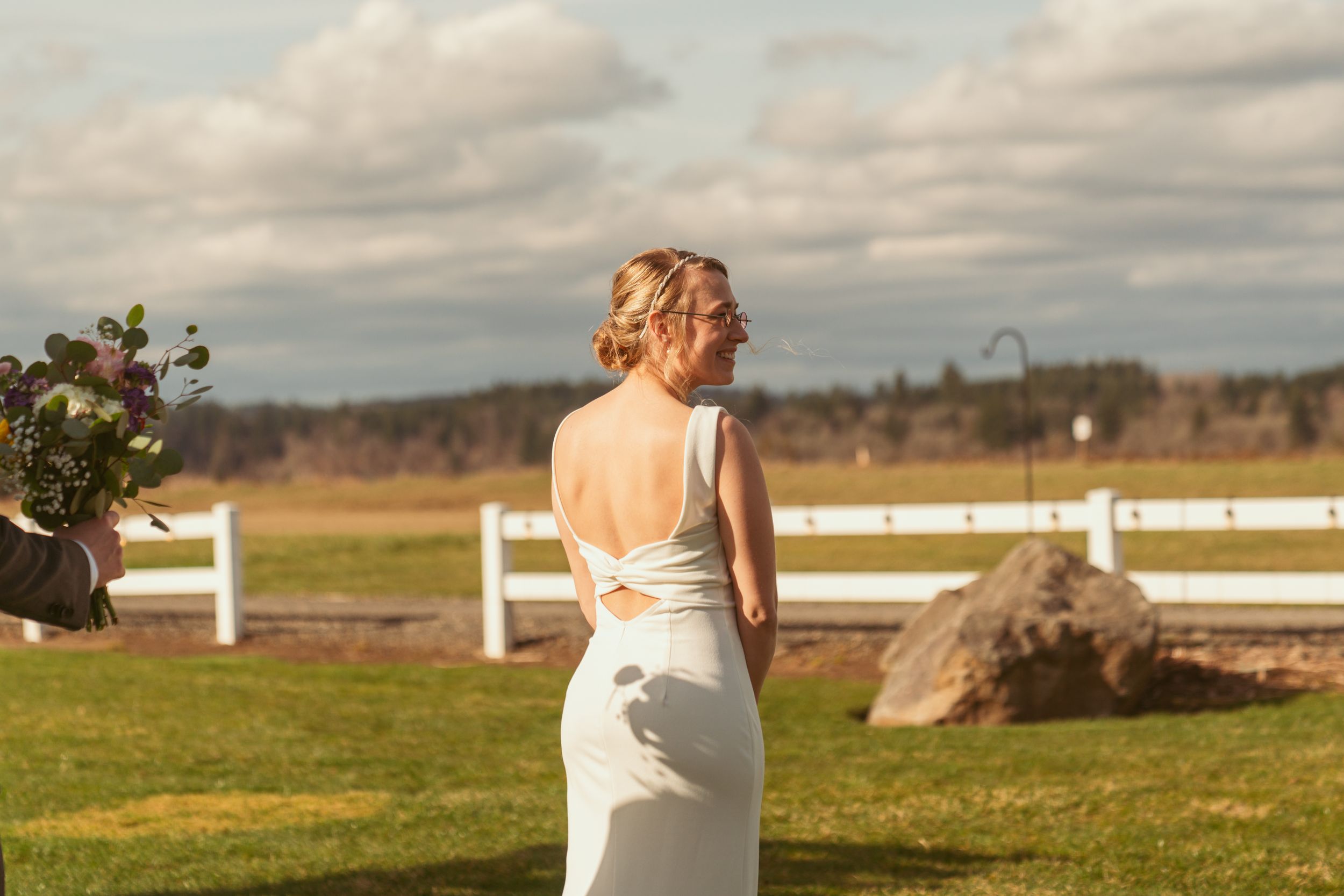A bride in a white wedding dress stands against a rustic countryside backdrop with white fences and open fields.