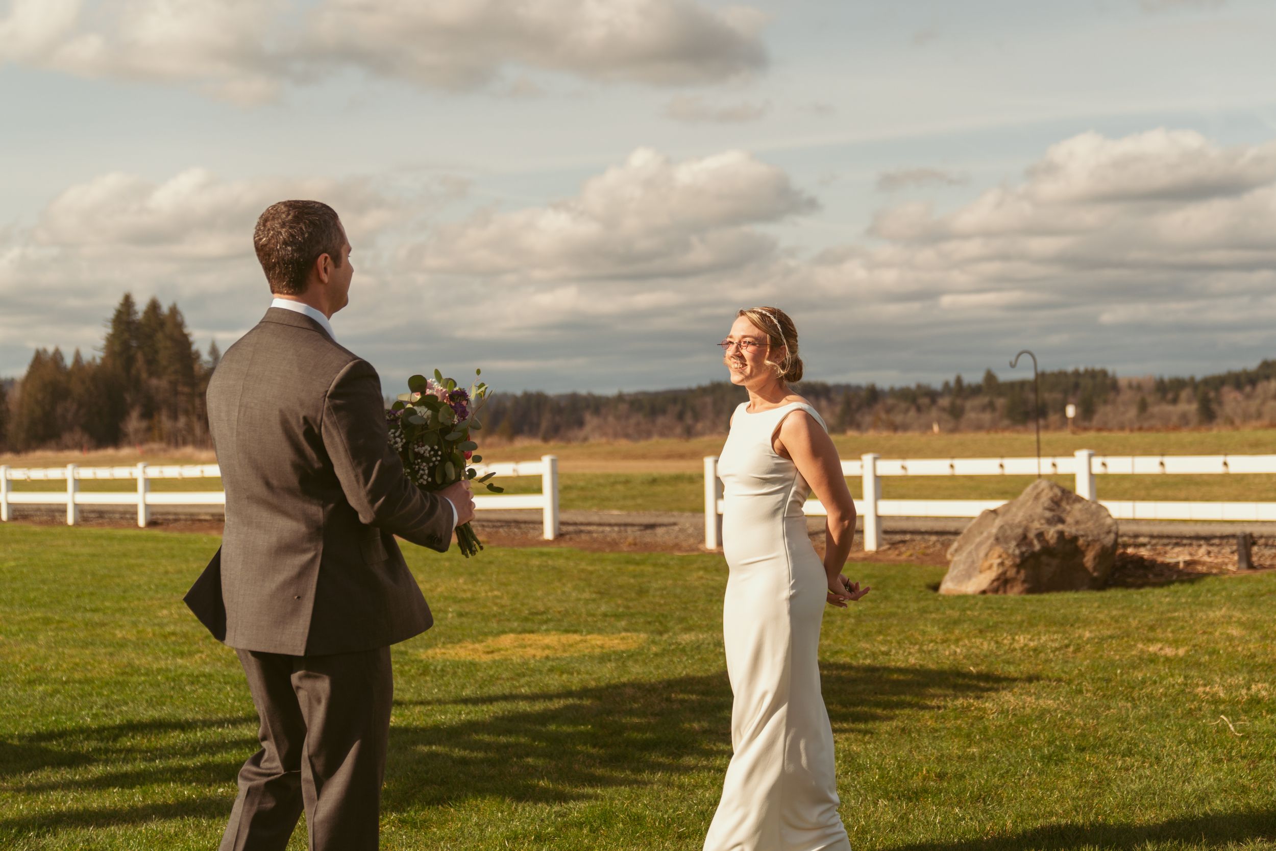 A couple shares a tender first look moment on their wedding day in a scenic field with white fences and cloudy skies.