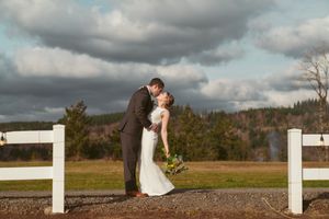 A romantic wedding couple embraces on a rustic country path bordered by white fences under dramatic cloudy skies.