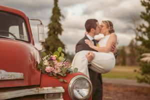 A romantic wedding couple embraces near a vintage red truck with a floral bouquet on the hood beneath cloudy skies.