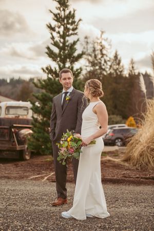 A bride and groom pose outdoors near pine trees and a vintage truck on a cloudy day.