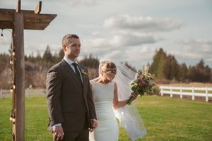 A couple in wedding attire stands together in a rustic outdoor setting with white fencing and cloudy skies.