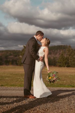 A romantic outdoor wedding moment captured on a scenic gravel path with dramatic cloudy skies in the background.