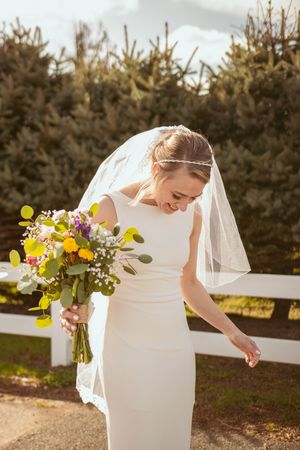 Elegant bride in white dress holding vibrant yellow and purple wildflower bouquet against evergreen backdrop.