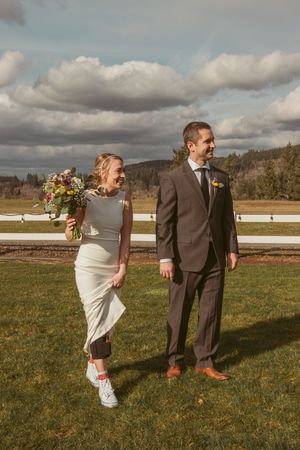 A playful outdoor wedding photoshoot in a grassy field with cloudy skies and white fence in background.